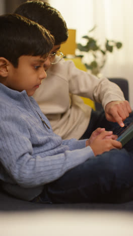 Vertical-Video-Of-Two-Young-Boys-Sitting-On-Sofa-At-Home-Playing-Games-Or-Streaming-Onto-Digital-Tablet-Together